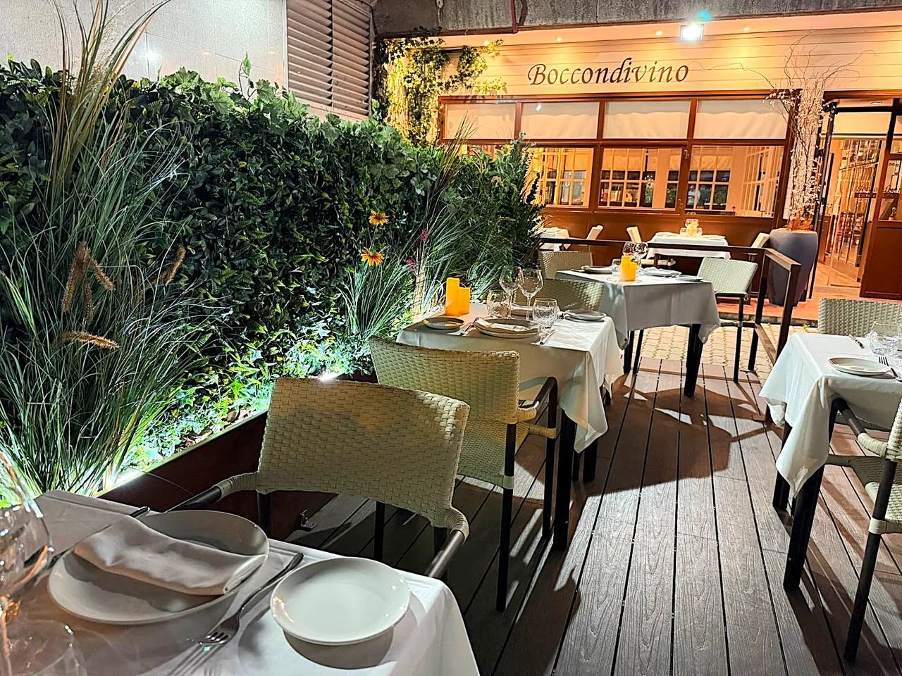 Terrace at night with white-linen tables laid on a wooden deck behind a tall green hedge, the lit Boccondivino sign and glazed dining room behind