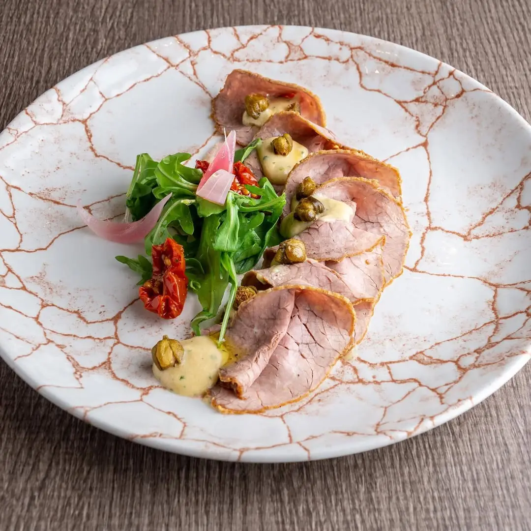 Sliced roast beef plated with arugula, sun-dried tomato, pickled onion and capers in mustard sauce