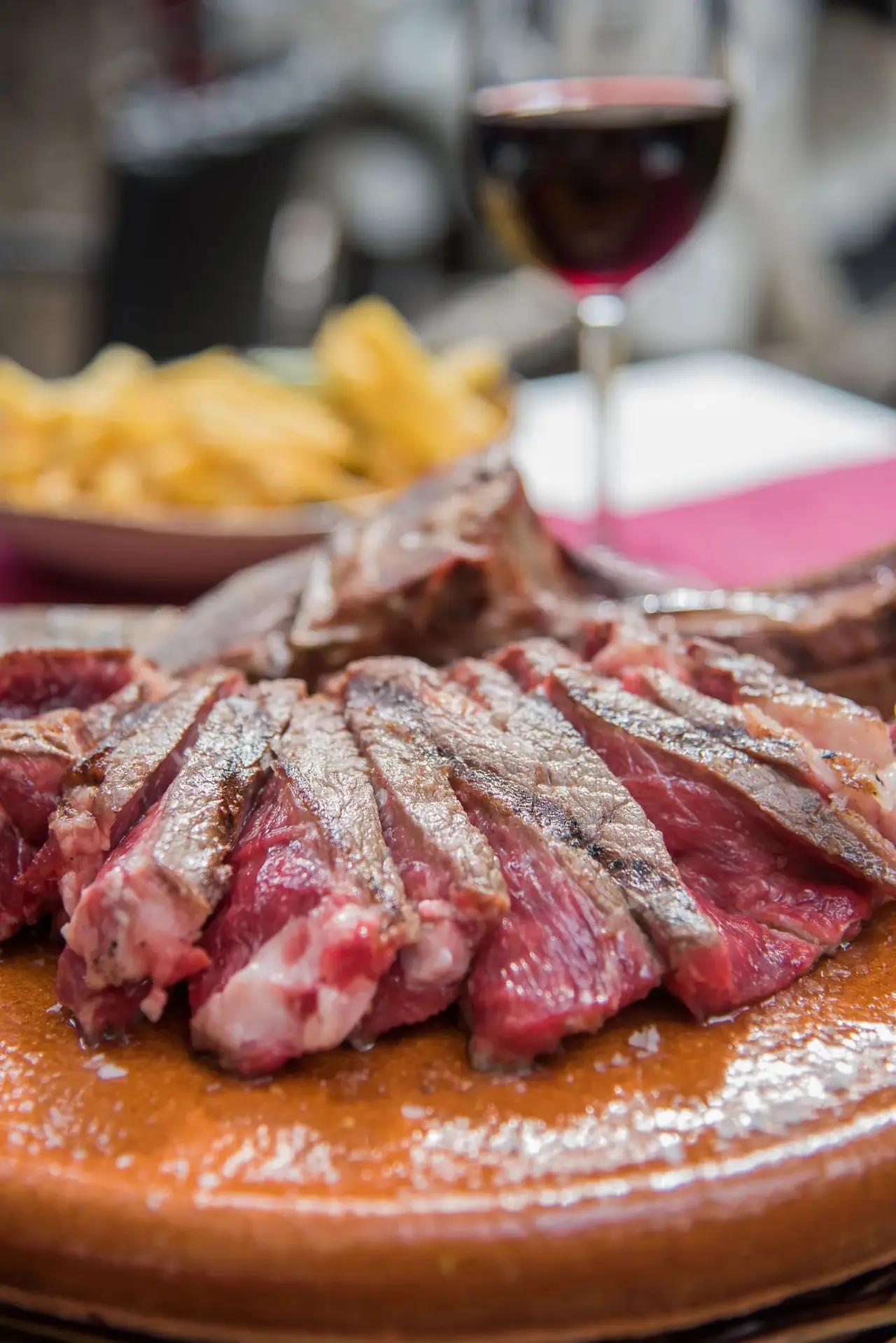 Sliced grilled steak fanned across a terracotta plate with a glass of red wine and a bowl of fries in the background, Bodega Joan