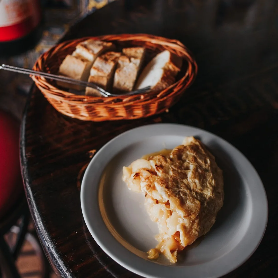 A slice of Spanish omelette on a white plate beside a basket of bread