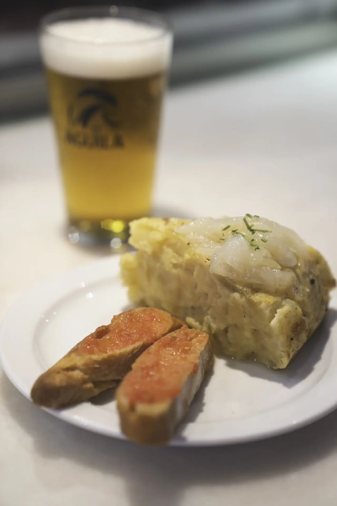A slice of Spanish potato tortilla topped with caramelized onion and herbs, served with two slices of pa amb tomaquet on a white plate, at Bodega La Peninsular