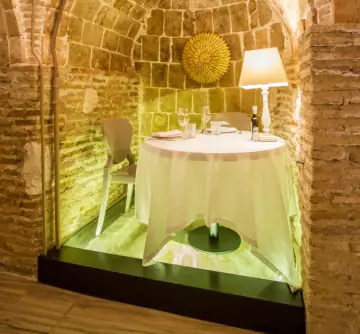 Round dressed table for two set in a stone-vaulted niche under green-tinted lighting with a lamp and sunburst wall ornament
