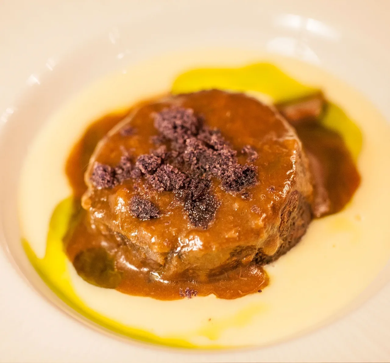 A round portion of braised meat topped with dark crumbs on a pool of yellow sauce