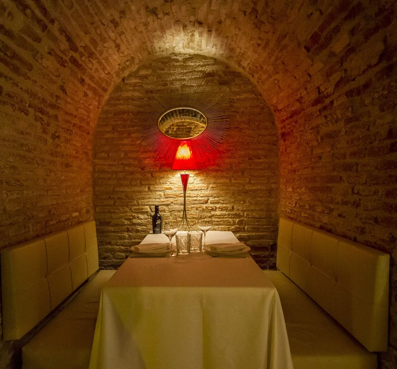 A small brick vaulted alcove with a table, cushioned benches and a red lamp on the wall