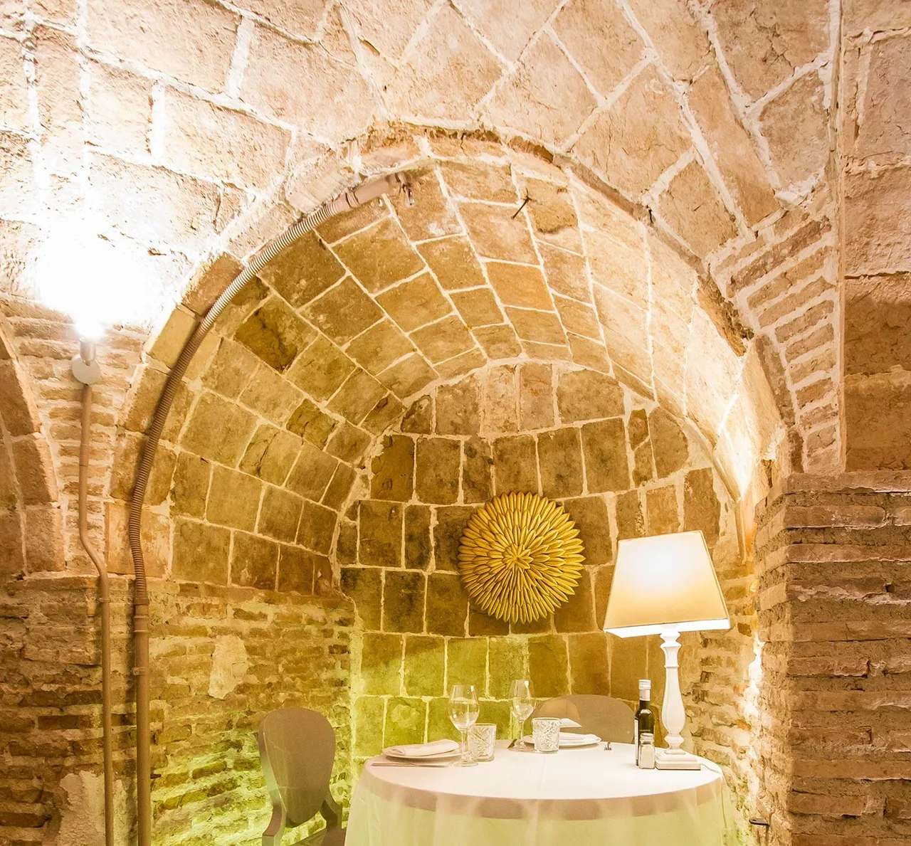 A round table set under a brick vault with a gold sunburst mirror and a lamp