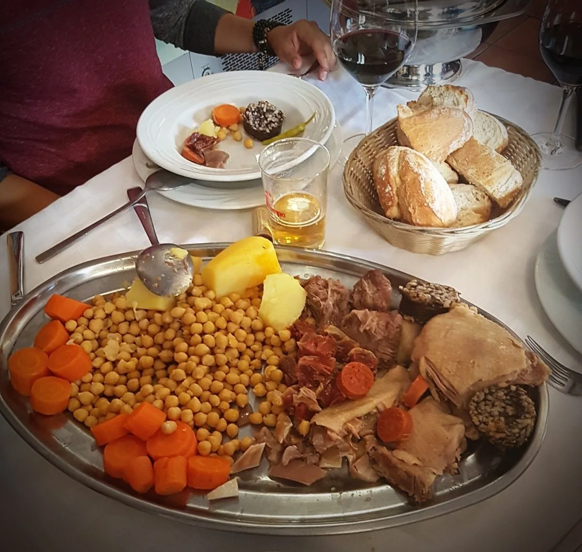 A steel platter of chickpeas, potatoes, carrots, meats and blood sausage beside a basket of bread