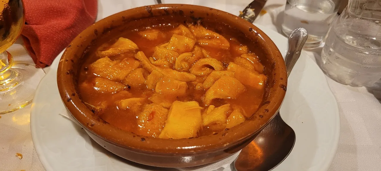 A clay cazuela of tripe stew in orange sauce with a spoon on a white plate