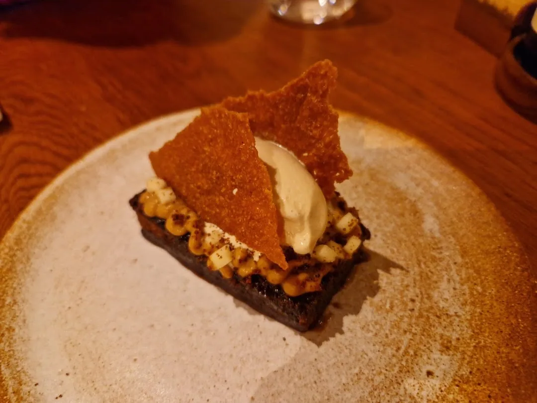 A dark charred bread base topped with corn, a crumble, a quenelle of cream, and a large crispy tuile shard