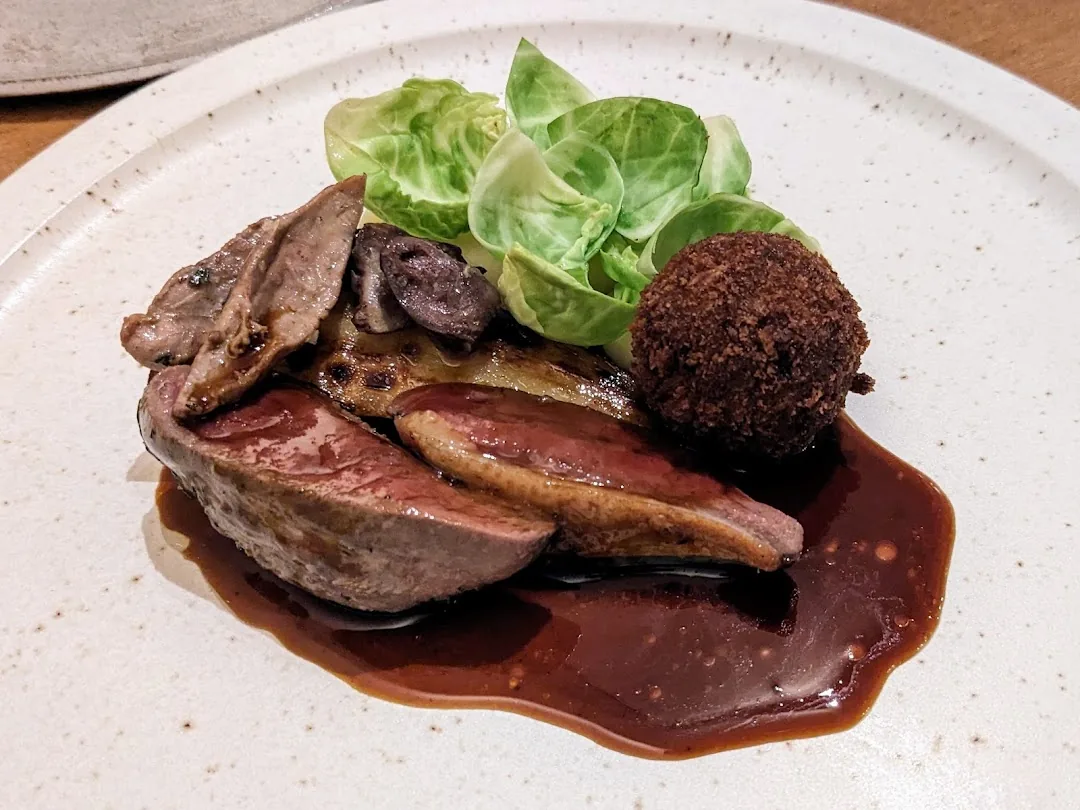 A plated meat dish with pink-seared slices, seared foie, a breaded croquette, roasted mushrooms, Brussels sprout leaves, and a dark glossy sauce