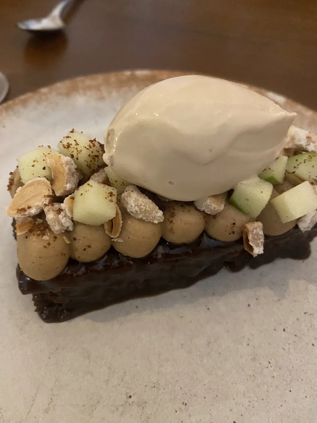 A layered chocolate dessert bar topped with peanuts, diced green apple, meringue pieces, and a quenelle of pale ice cream