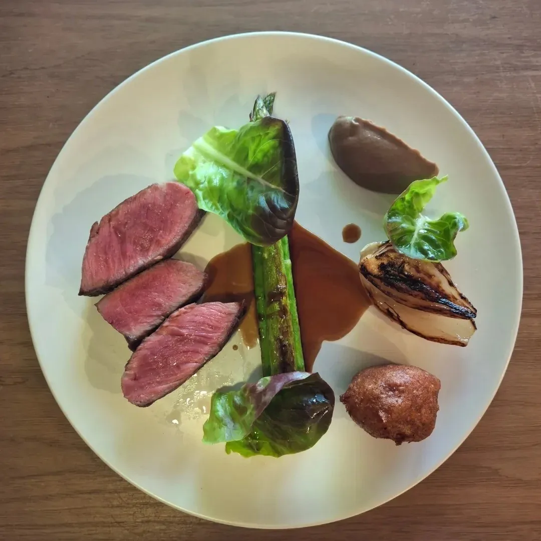 Slices of pink roast meat, a green stalk, an onion and two dark round pieces on a large white plate