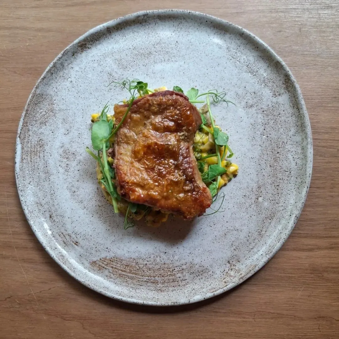 A browned piece of meat on a grey speckled plate with a corn mixture and green shoots