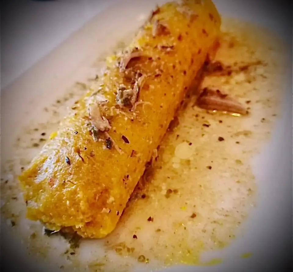 A golden cornmeal roll topped with shredded meat and herbs sitting in a pale sauce on a white plate