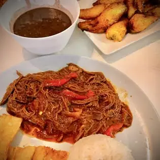 Plate of shredded beef in red sauce with red peppers, white rice and fried plantains, a bowl of dark sauce and fried plantains behind