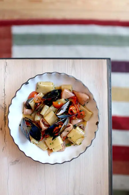 Overhead shot of a white ceramic bowl with mussels, pasta, and cherry tomatoes in broth on a wooden table, Bosco de Lobos.