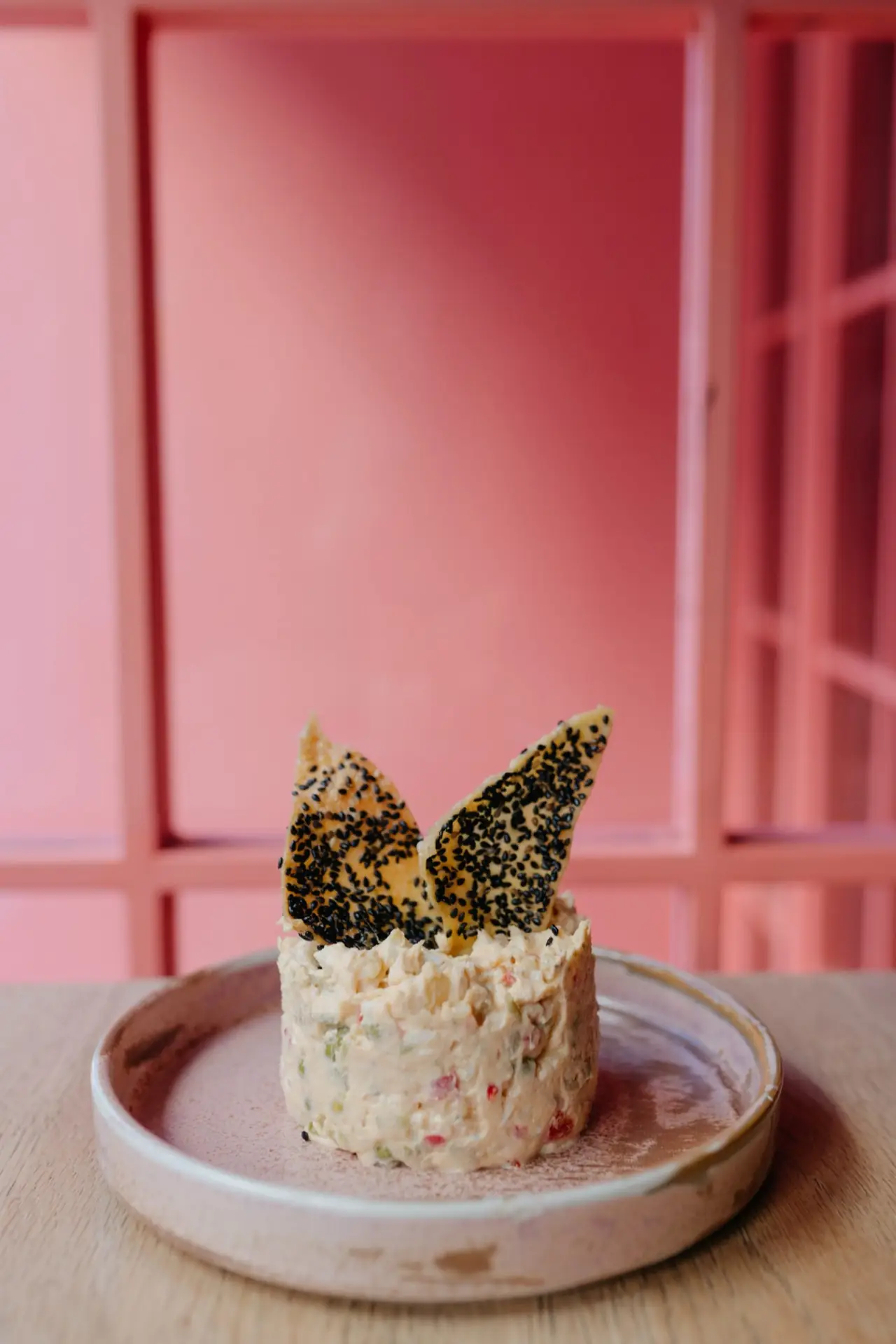 A molded creamy salad topped with two black-sesame tuile crackers on a rustic ceramic plate dusted with spice, against a pink panelled wall