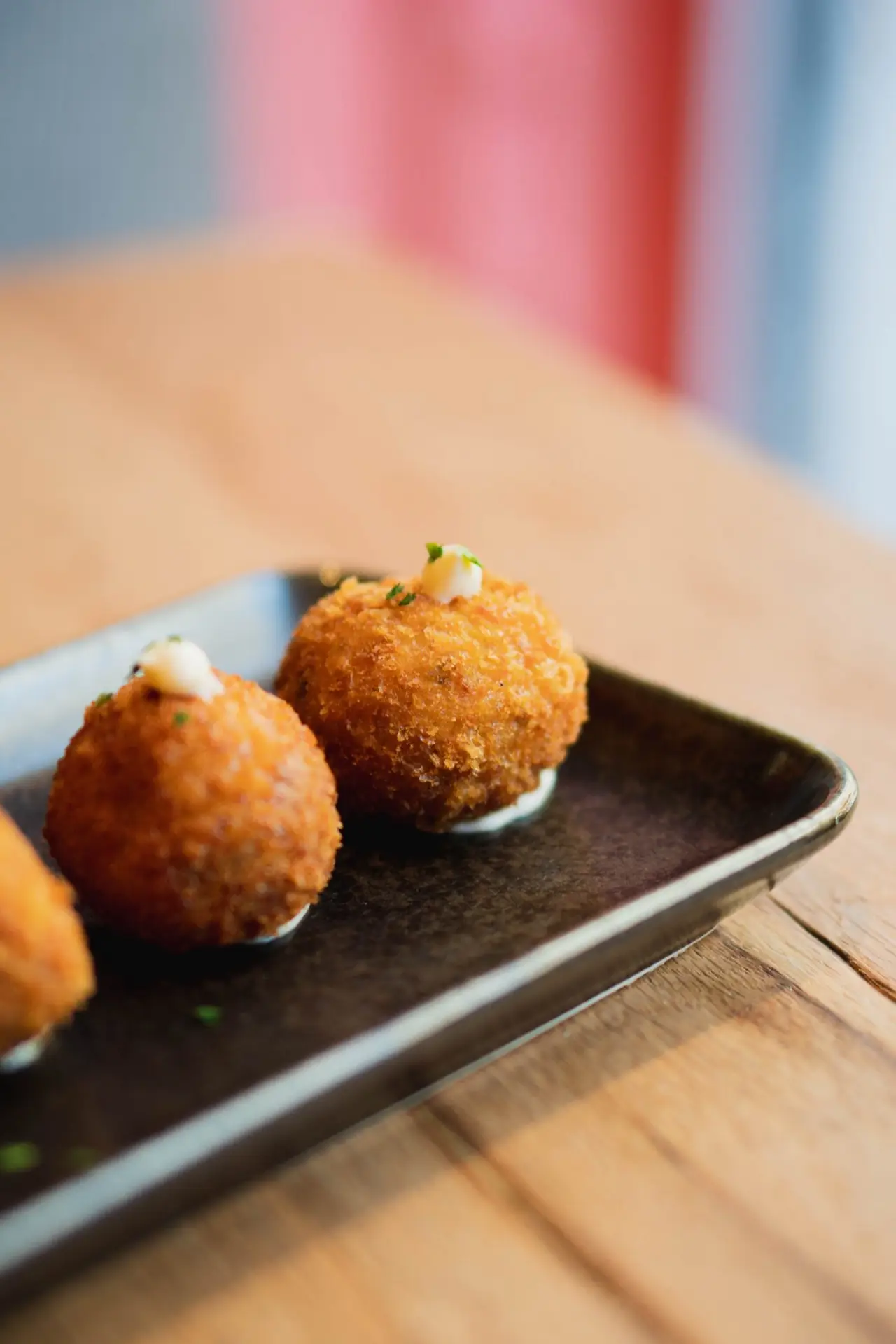Two golden breaded croquette balls each topped with a dot of aioli on a dark rectangular plate, on a wood table