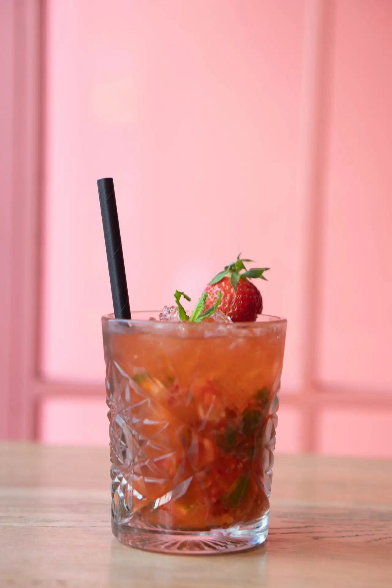 A strawberry and mint cocktail over crushed ice in a cut-glass tumbler with a black straw, garnished with a whole strawberry, against a pink wall