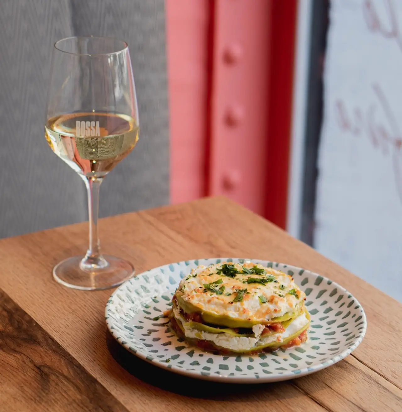 A layered courgette and cheese stack topped with melted cheese and chopped herbs on a green-flecked plate, beside a BOSSA-etched glass of white wine