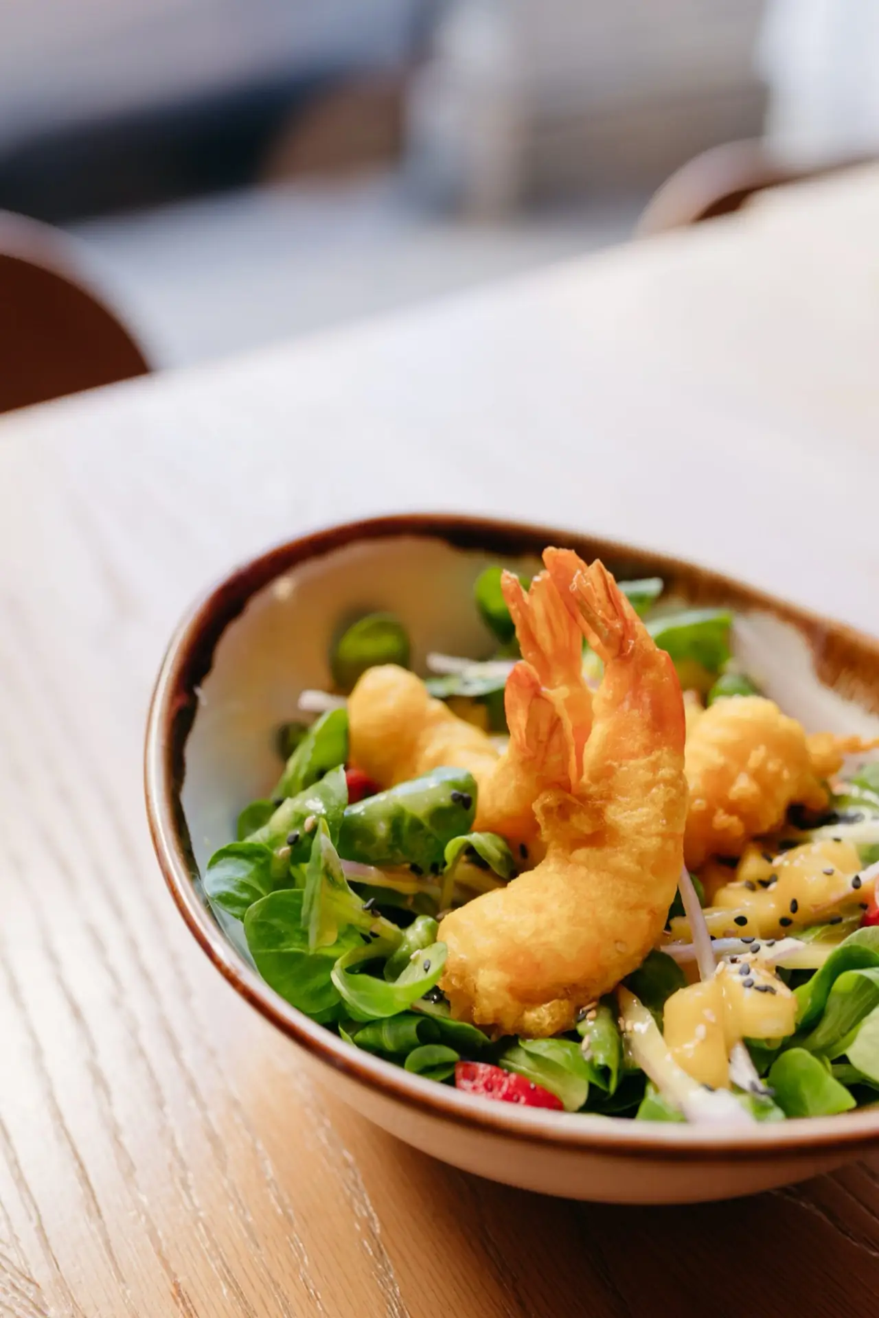 Tempura prawns over a lamb's lettuce salad with sesame seeds, sprouts and mango dressing in a brown-rimmed bowl on a wood table
