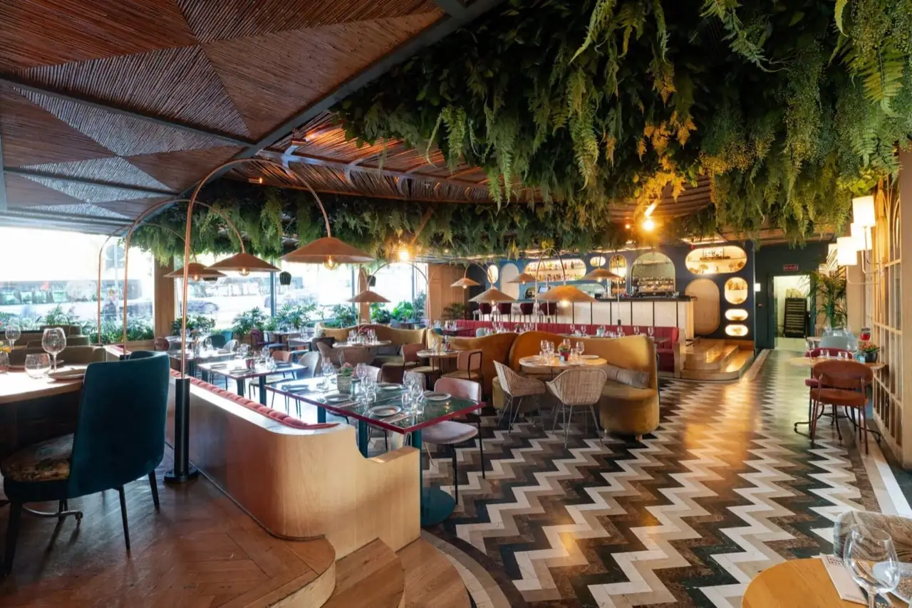 Lush restaurant dining room with hanging ferns covering the bamboo ceiling, black-and-white chevron tile floor, and mixed velvet seating.