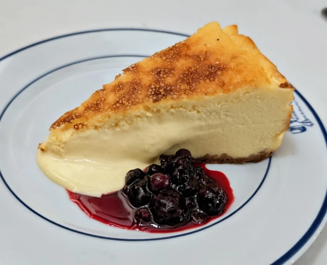 A slice of baked cheesecake with a caramelised top on a blue-rimmed plate with a berry compote