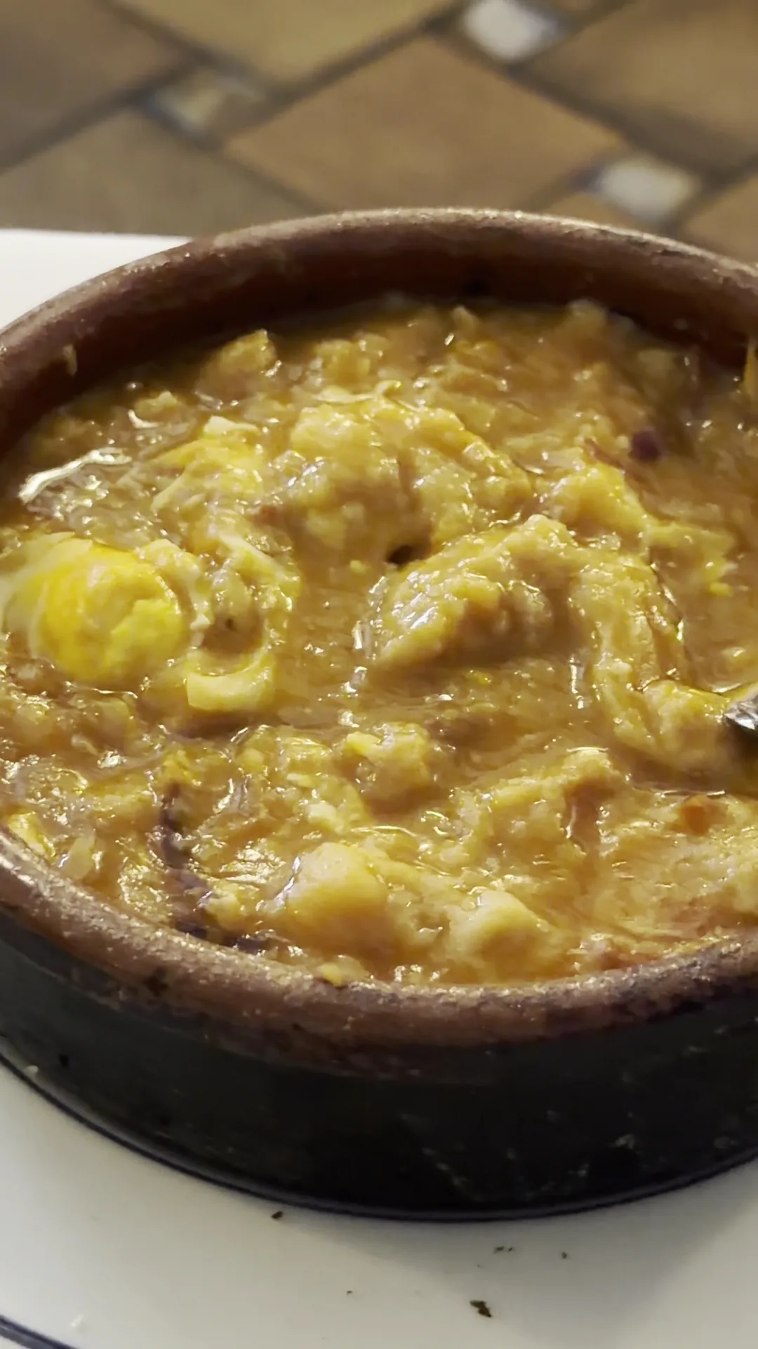 A rustic clay dish of scrambled eggs mixed with browned chunks of meat, glistening with oil, on a white cloth