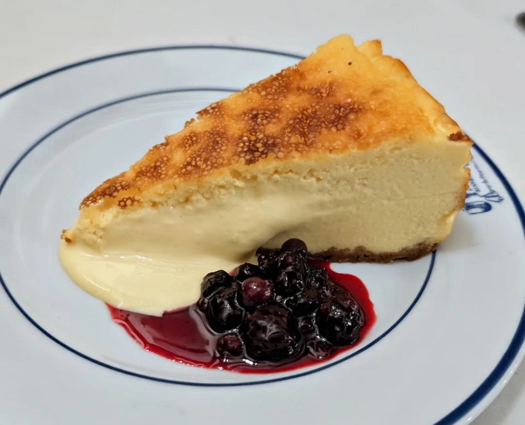A slice of baked cheesecake with a caramelized top on a blue-rimmed plate, with a swirl of cream and a blueberry compote