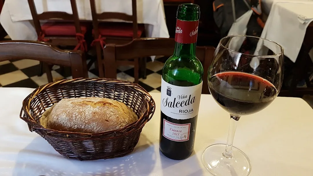 A basket of crusty round bread beside an open bottle of Rioja red wine and a full glass of red wine on a white tablecloth