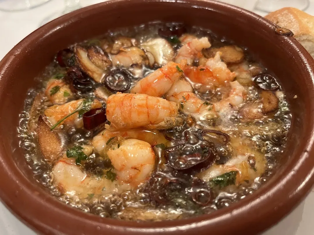 Garlic shrimp sizzling in oil in a brown clay dish, garnished with parsley and chili, with a piece of bread at the edge