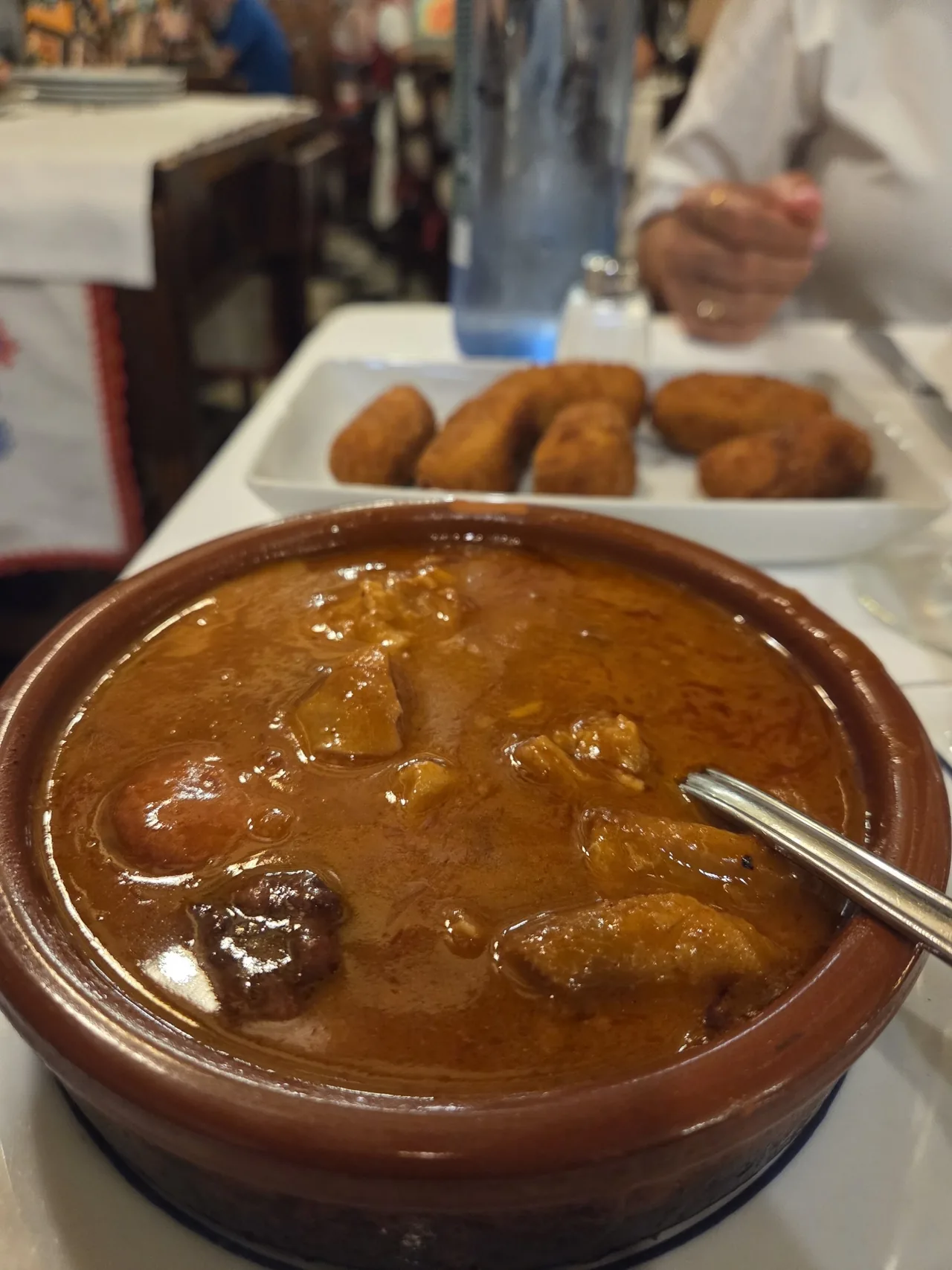 A clay cazuela of tripe stew in a dark orange sauce with a fork, and a plate of croquetas behind