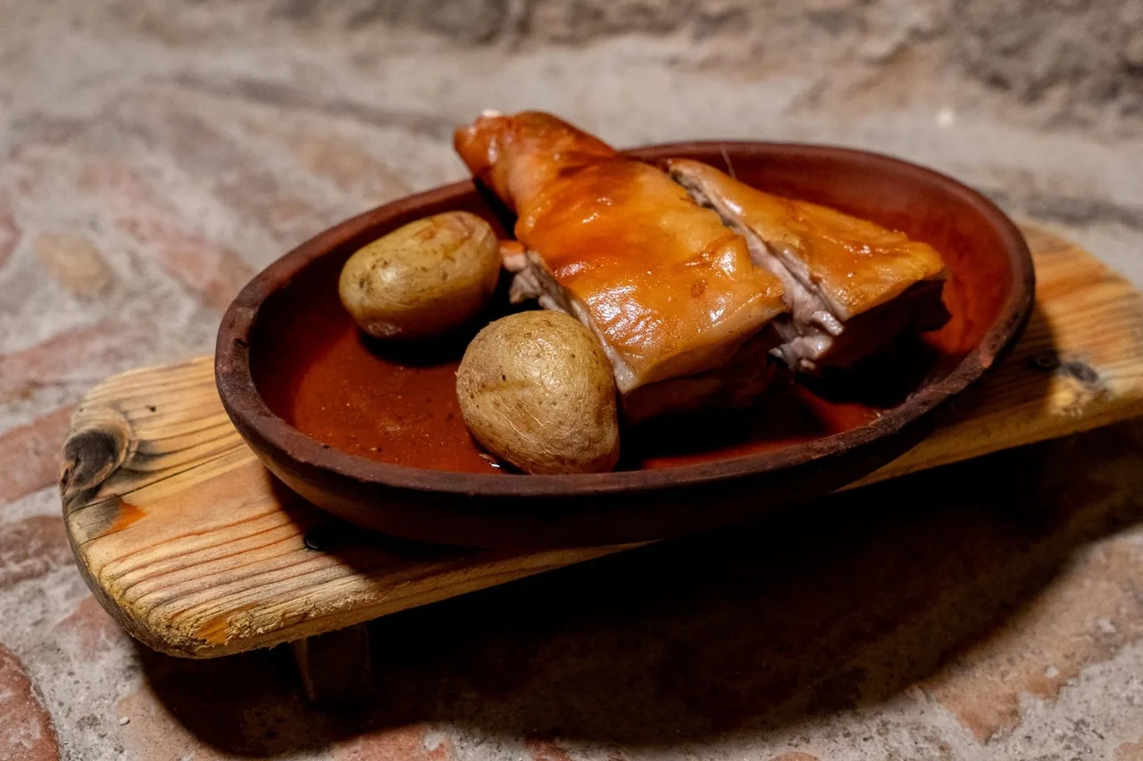 A portion of roast suckling pig with two boiled potatoes in a clay dish on a wooden board