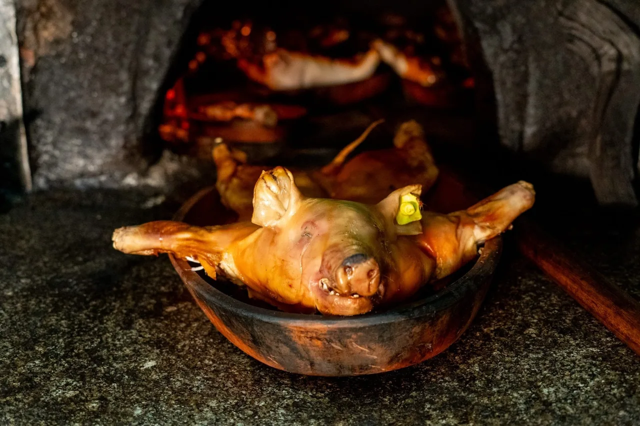 A whole roast suckling pig in a clay dish in front of a wood-fired oven