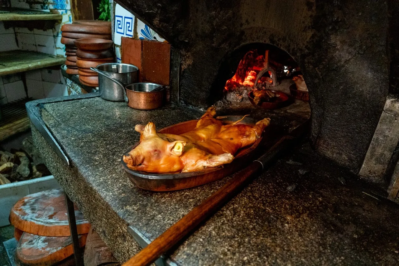 A whole roast suckling pig on a tray beside a stone counter and a brick oven with glowing embers