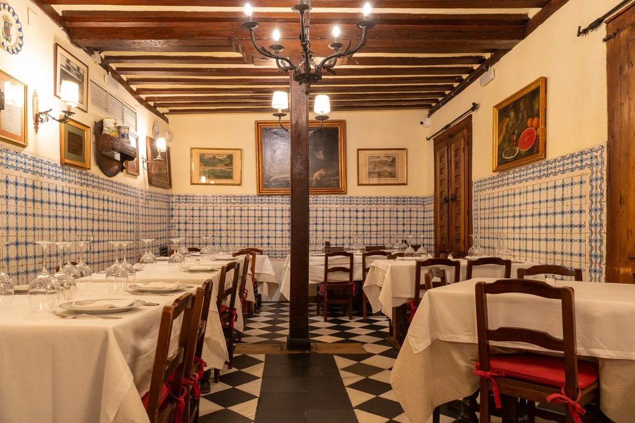 A dining room with exposed wooden ceiling beams, blue and white tiled walls, framed paintings and a black and white checkered floor.