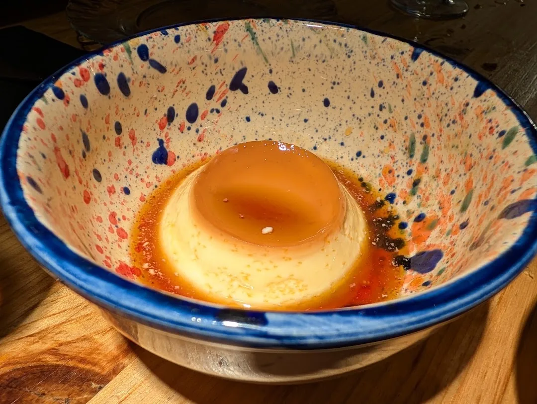 A flan dessert in caramel sauce, served in a hand-painted blue and orange speckled bowl