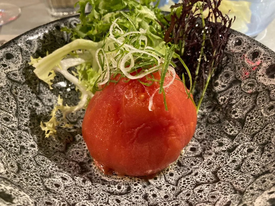 A peeled whole tomato topped with shaved fennel and mixed leafy greens on a textured black plate