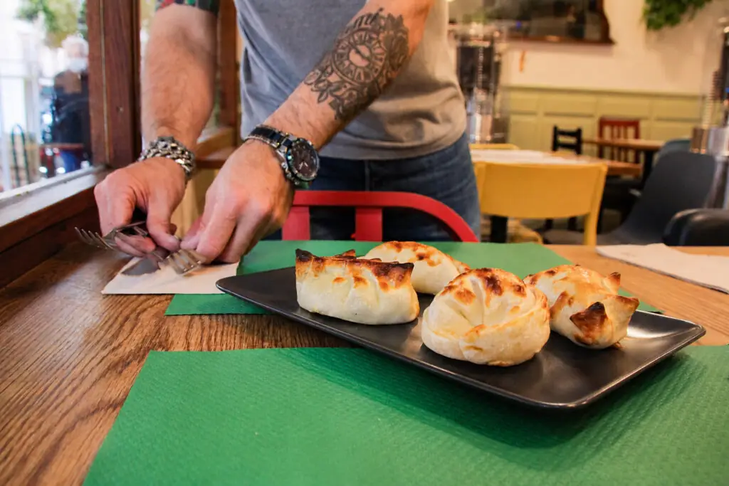 Three baked empanadas on a black rectangular plate on a dining table, a server setting cutlery nearby with the dining room behind