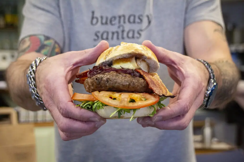 Burger with a beef patty, melted cheese, bacon, caramelised onion and tomato held in two hands