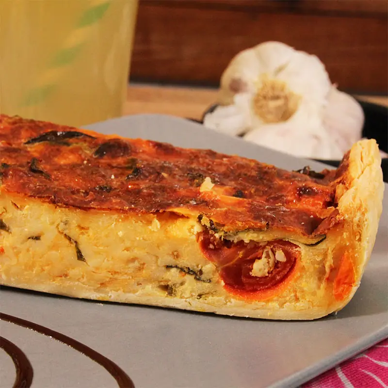 Slice of quiche topped with tomato, served beside a head of garlic.
