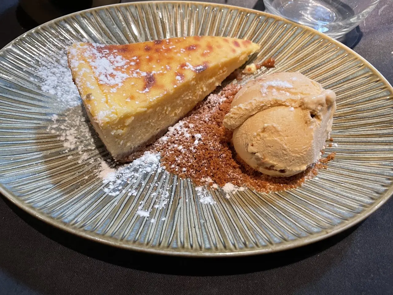 A slice of cheesecake on a plate with a scoop of ice cream and a dusting of powdered sugar.
