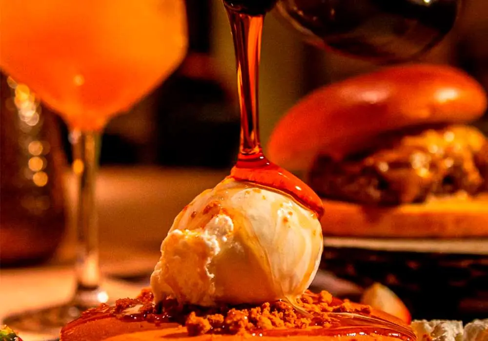 Caramel sauce being poured over a scoop of ice cream on a dessert plate, with a cocktail and burger in the background.