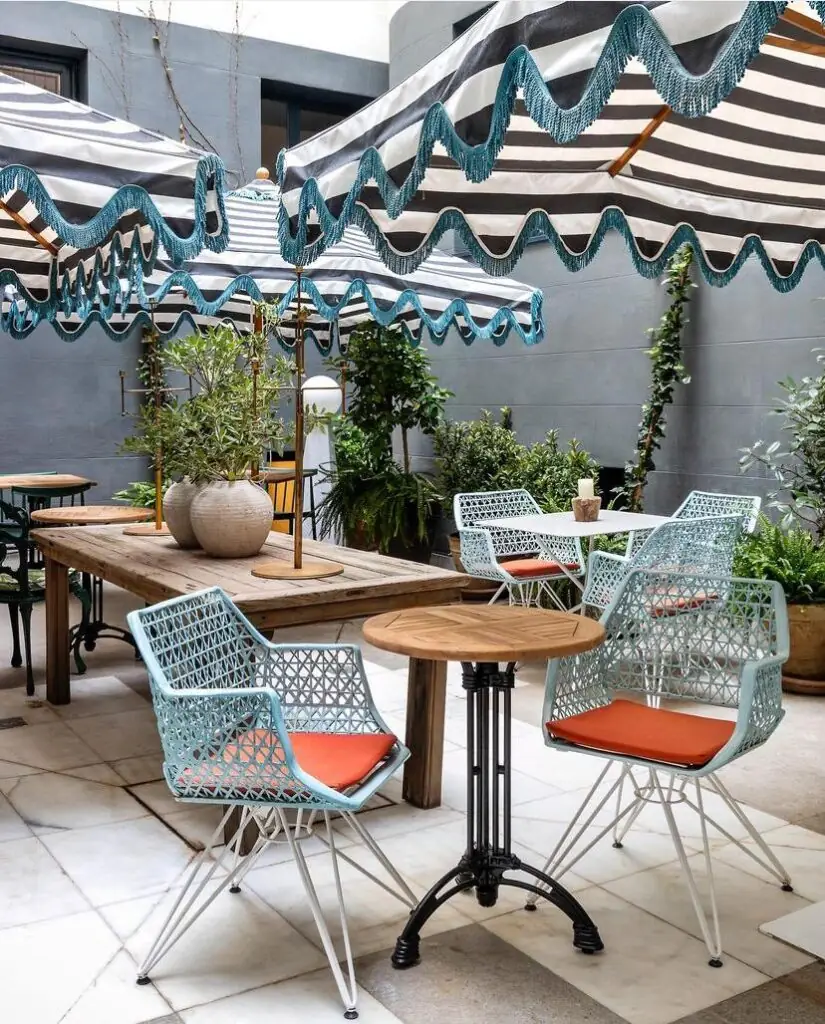 Outdoor terrace with striped fringed parasols, pale blue lattice chairs with orange cushions, wooden tables and potted greenery against a grey wall