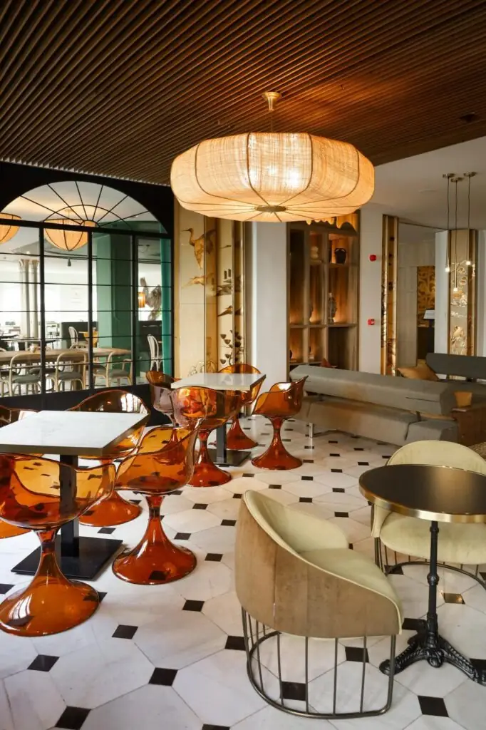 Interior lounge with amber acrylic tulip chairs, a woven pendant lamp, velvet armchairs and a decorative crane-motif screen on black-and-white tiled floor