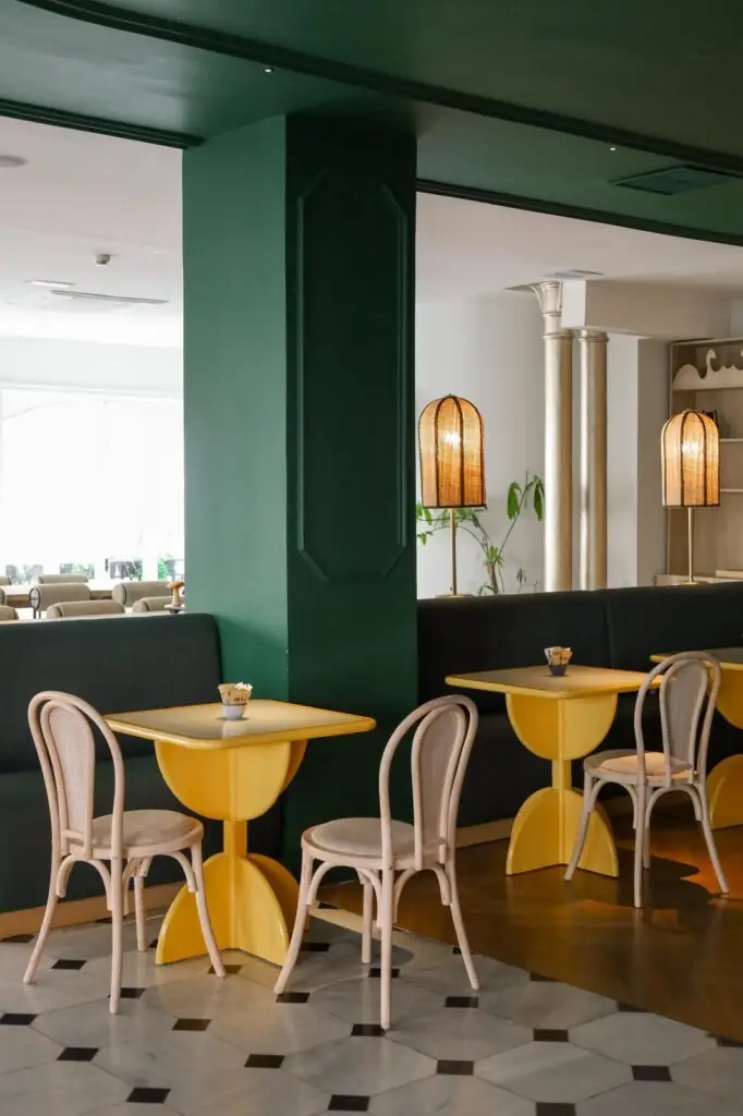 Dining nook with a green painted column, yellow pedestal tables, pale cane bistro chairs and rattan table lamps on chequered tile floor