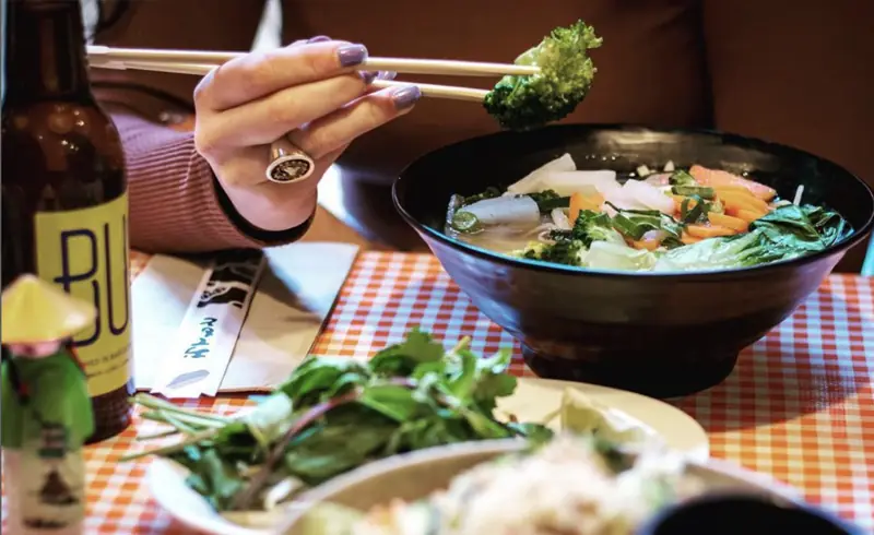 Bowl of Vietnamese pho with herbs, carrot, radish, and greens, a hand lifting a piece of broccoli with chopsticks, at Bun Bo