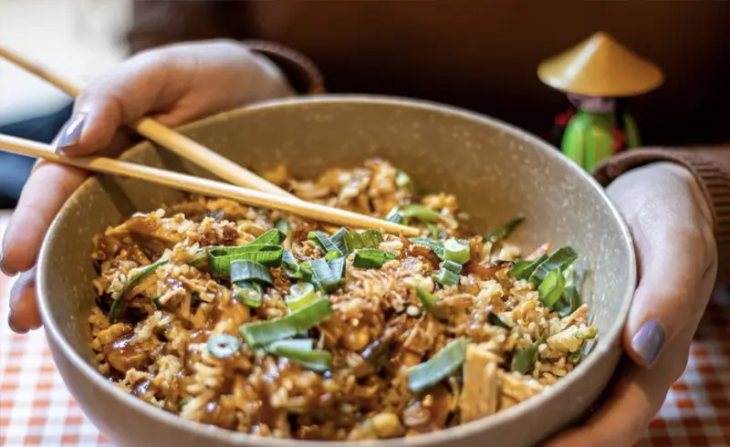 Bowl of fried rice with scallions and tofu held with chopsticks resting on top, at Bun Bo