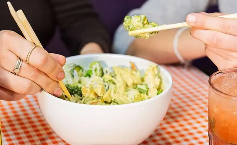 White bowl of noodle salad with broccoli and battered chicken, chopsticks lifting a piece of broccoli in tempura batter, at Bun Bo