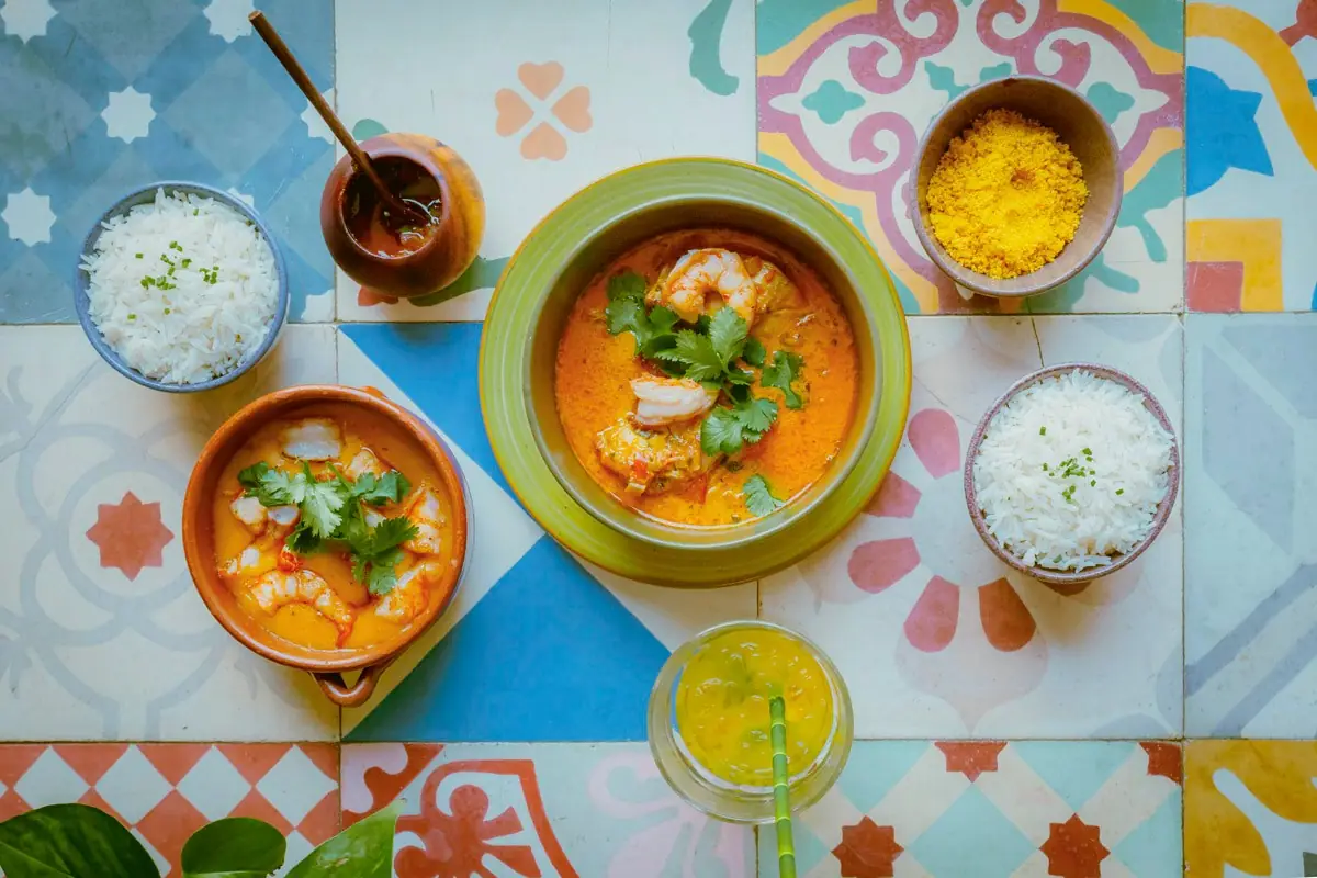 Overhead shot of Brazilian-style seafood moqueca stews with shrimp and cilantro, served alongside bowls of white rice and a citrus drink, on a colorful patterned tile tabletop at Buriti