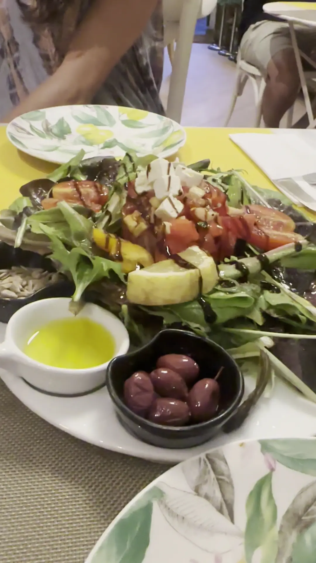 A green salad with tomatoes, courgette, and olives, served with a small bowl of olive oil.